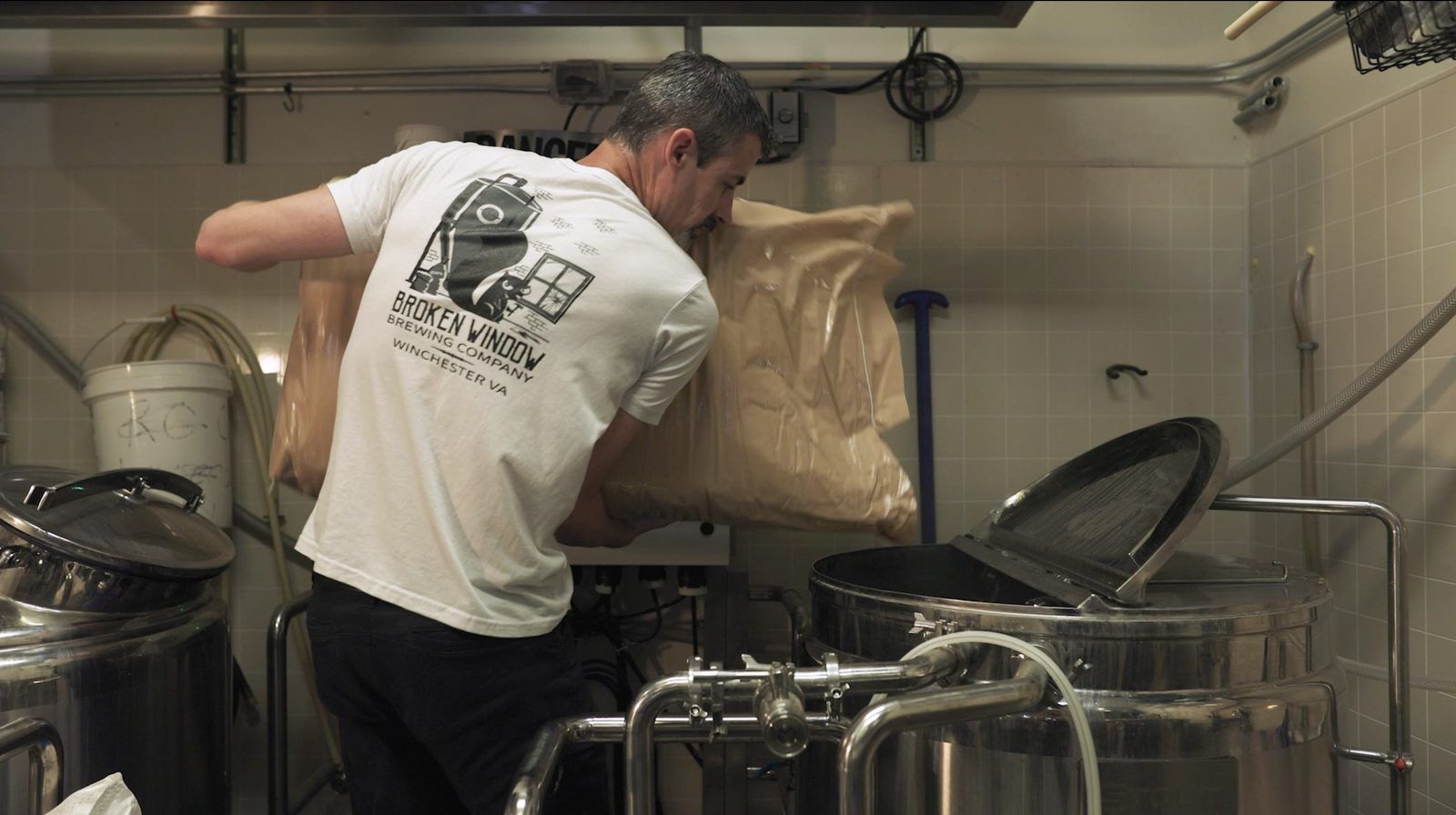 A brewer in a Broken Window Brewing Company T-shirt pours a sack of grain into the brew kettle.